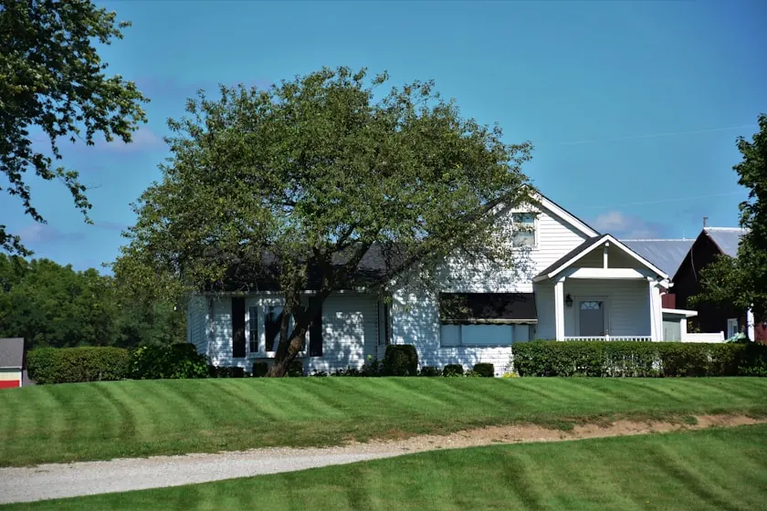 A white house with a gabled porch under a broad shade tree in Coweta, with a striped mown lawn running down to a gravel drive and a red barn roof behind